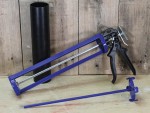 A purple caulking gun with a black handle is lying on a wooden surface next to a black cylindrical tube and a long, thin rod with a handle, all set against a wooden plank background.