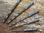 Five metal drill bits of different sizes are arranged on a textured, brownish surface resembling stone or wood grain. The drill bits have hexagonal shanks and sit parallel to each other.