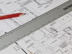 A close-up of architectural blueprints with a metal ruler and a red pencil placed on top, showing detailed building plans, measurements, and room layouts.
