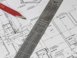 A metal ruler with measurement conversions and a red pencil rest on detailed architectural blueprints, showing floor plans and room layouts for a building.