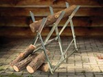 A metal sawhorse holds a large log, with several cut logs lying on the ground beneath it. The scene is set on a stone-paved surface with a wooden wall in the background.