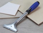 A heavy-duty scraper with a blue and black handle rests on a wooden surface next to beige ceramic tiles and a white tile.,