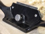 Close-up of a black metal spokeshave tool with a shiny adjustment knob and blade, resting on a light surface with wood shavings in the background.