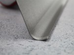 Close-up of the edge of a metal spatula resting on a light gray textured surface. The focus is on the smooth, beveled metal edge.