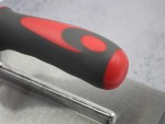 Close-up of a hand tool with a black and red ergonomic handle attached to a flat metal surface, likely a trowel, on a light grey background.
