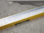 Close-up of a metallic edge with fine, parallel lines running along its length, attached to a yellow surface, resting on a textured, light gray background.