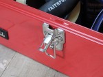 Close-up of a silver latch on a red metal toolbox, resting on a wooden surface, with various tools and supplies visible inside the box.