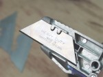 A close-up of a utility knife with a metal blade engraved HEAVY DUTY MADE IN SHEFFIELD ENGLAND. Two spare blades are visible in the blurred background on a wooden surface.,