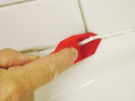 A hand uses a red plastic tool to apply or smooth white caulk along the seam between white bathroom tiles and a bathtub.,