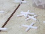 White plastic tile spacers are scattered on a beige tiled surface, with one spacer partially placed in the gap between two tiles. A plastic container with more spacers is visible in the background.
